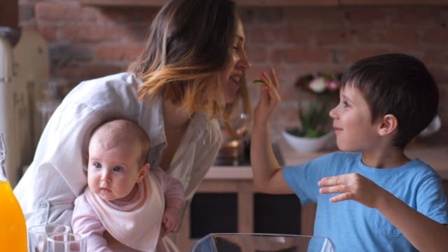Una mujer sostiene a un bebé mientras interactúa con un niño pequeño en una cocina. El bebé lleva un babero y el niño lleva una camisa azul. Se ve una botella de jugo de naranja en el mostrador.