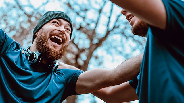 Dos personas están al aire libre con ropa deportiva. Una lleva un gorro y auriculares y sonríe ampliamente. Los árboles se ven borrosos al fondo.
