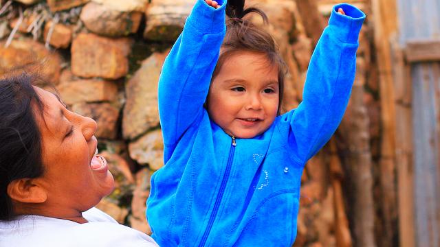 Una mujer levanta a un niño sonriente con una chaqueta azul, con un muro de piedra al fondo.