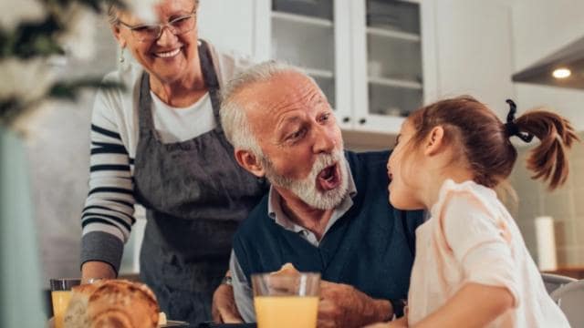 Un hombre mayor y una niña comparten un momento de juego en una mesa de comedor, mientras una mujer mayor está de pie cerca, sonriendo. Sobre la mesa hay una hogaza de pan y vasos de jugo.