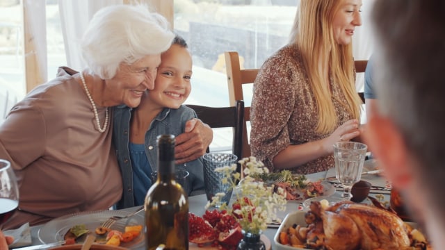 Una mujer mayor abraza a un niño sonriente en una mesa de comedor con varios platos. Otras personas también están sentadas y comiendo.