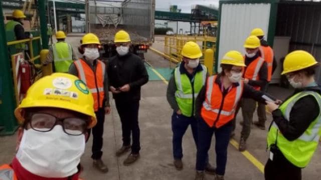 Un grupo de trabajadores de la construcción que visten cascos amarillos, máscaras faciales y chalecos reflectantes se encuentran juntos en un sitio de trabajo.