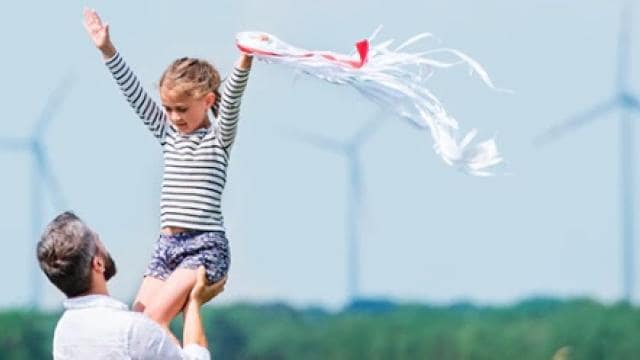 Un hombre levanta a un niño en el aire. El niño sostiene una cometa. Al fondo se ven turbinas eólicas.