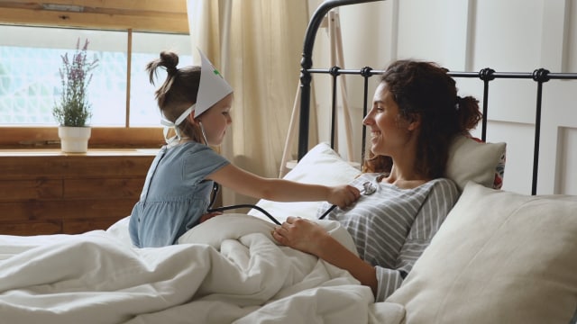 Un niño con un gorro de enfermera utiliza un estetoscopio de juguete para mirar a una mujer acostada en la cama. Ambos sonríen. Al fondo hay una ventana con plantas.