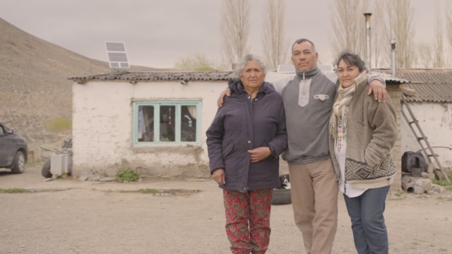 Tres personas se encuentran frente a una pequeña casa rústica con paneles solares en el techo. Al fondo se ve un paisaje árido y árboles sin hojas.