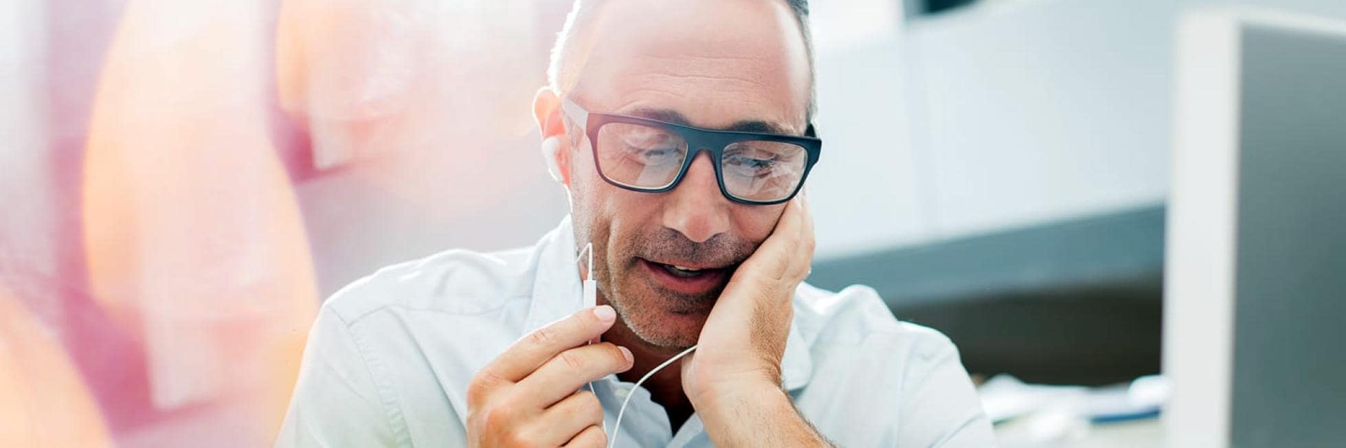 Un hombre con gafas y auriculares mantiene una conversación, tocándose la cara con la mano, en un entorno de oficina luminoso.