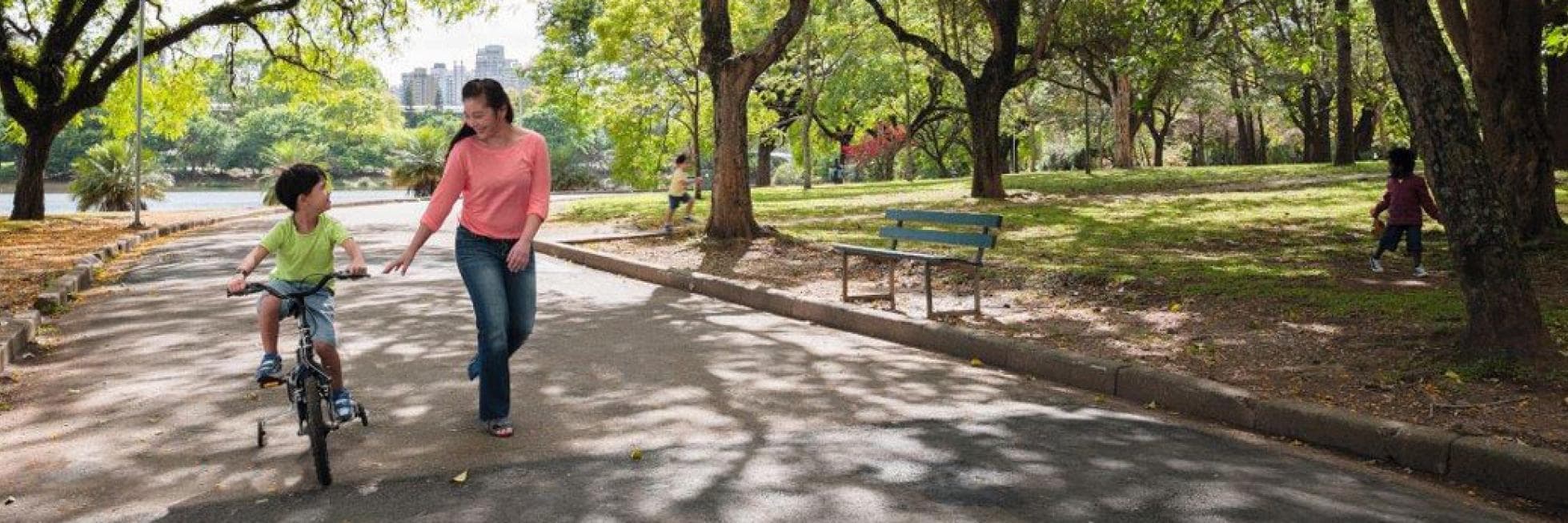 Una mujer camina junto a un niño que está aprendiendo a andar en bicicleta en un sendero del parque, rodeado de árboles y césped verde.