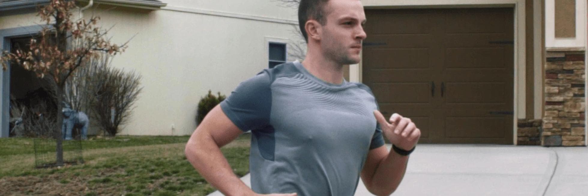 Persona haciendo jogging en una calle residencial, vistiendo una camiseta gris, con casas y un camino de entrada al fondo.