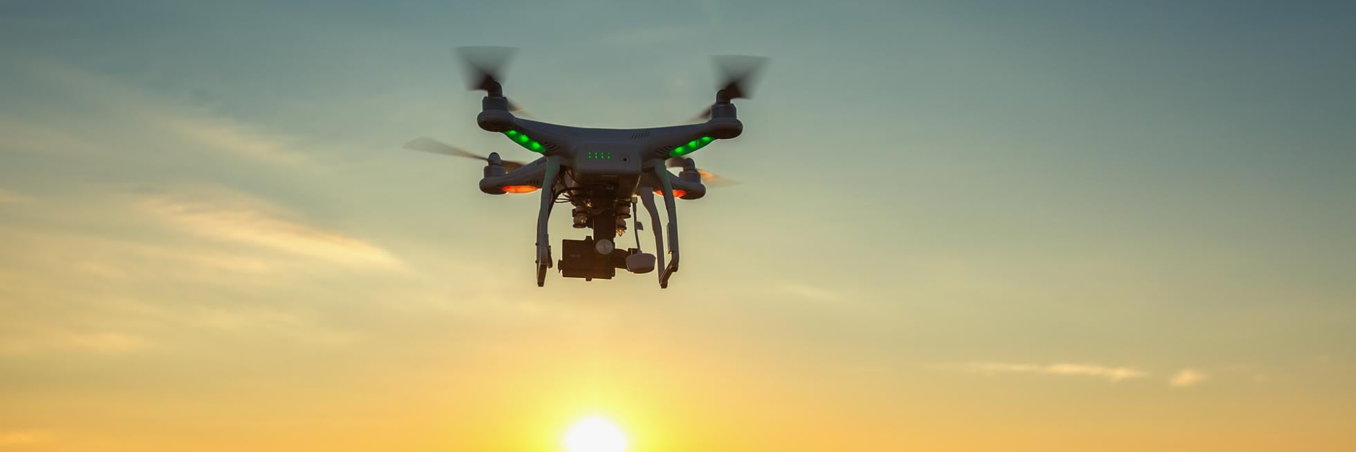 Un dron vuela sobre un campo al atardecer, con el sol iluminando el cielo y el paisaje.