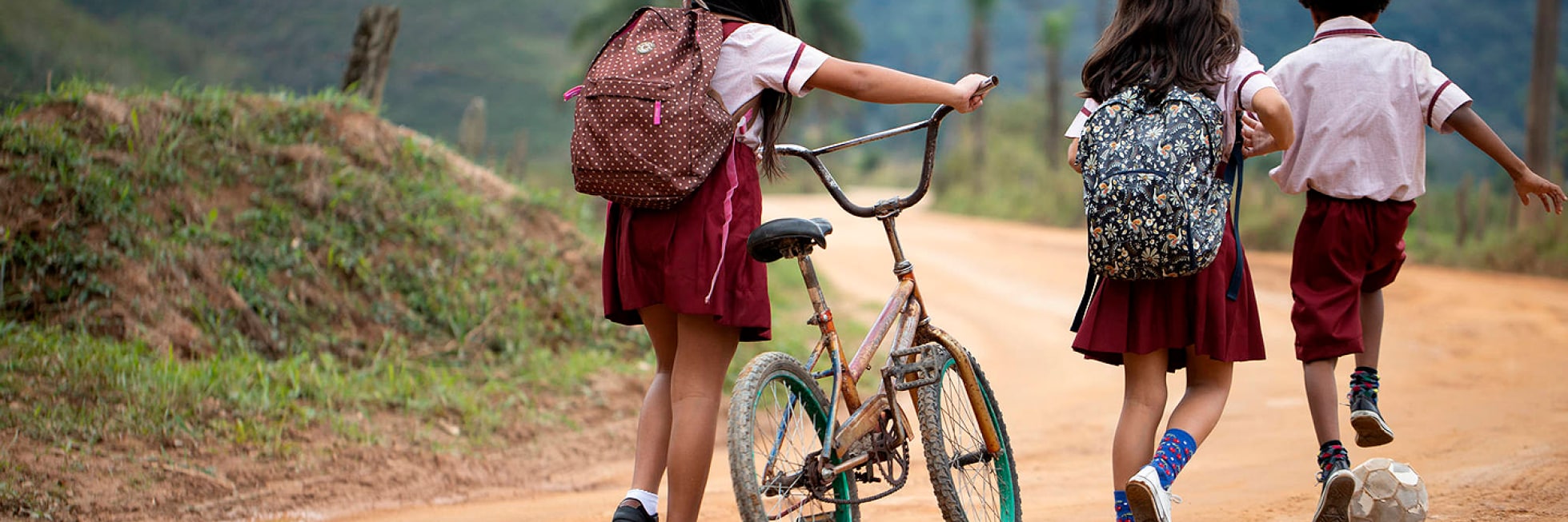 Tres niños con uniformes escolares caminan por un camino de tierra; dos llevan mochilas y uno empuja una bicicleta.