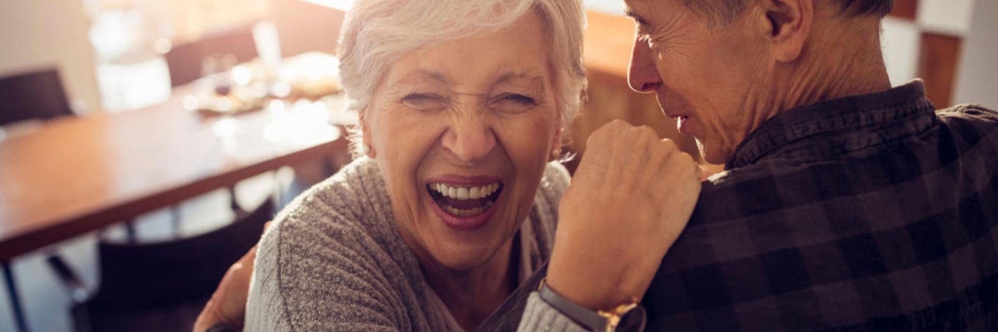 Una anciana ríe alegremente mientras abraza a un hombre en el interior, con una mesa y sillas de madera al fondo.