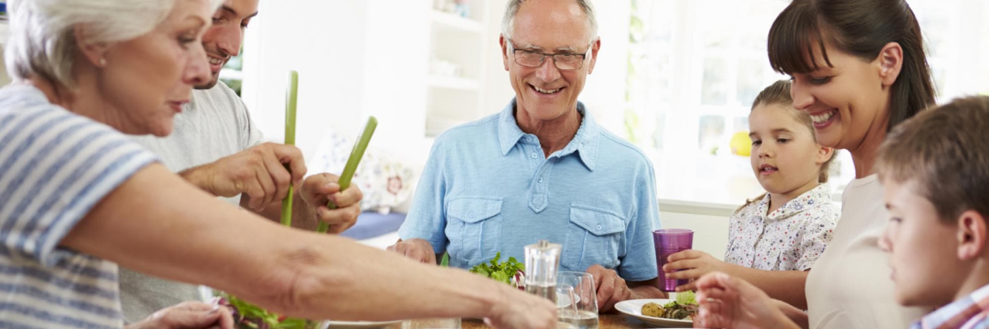 Una familia disfruta de una comida junta en una mesa de comedor, sonriendo y sirviendo comida. El ambiente es luminoso y alegre.