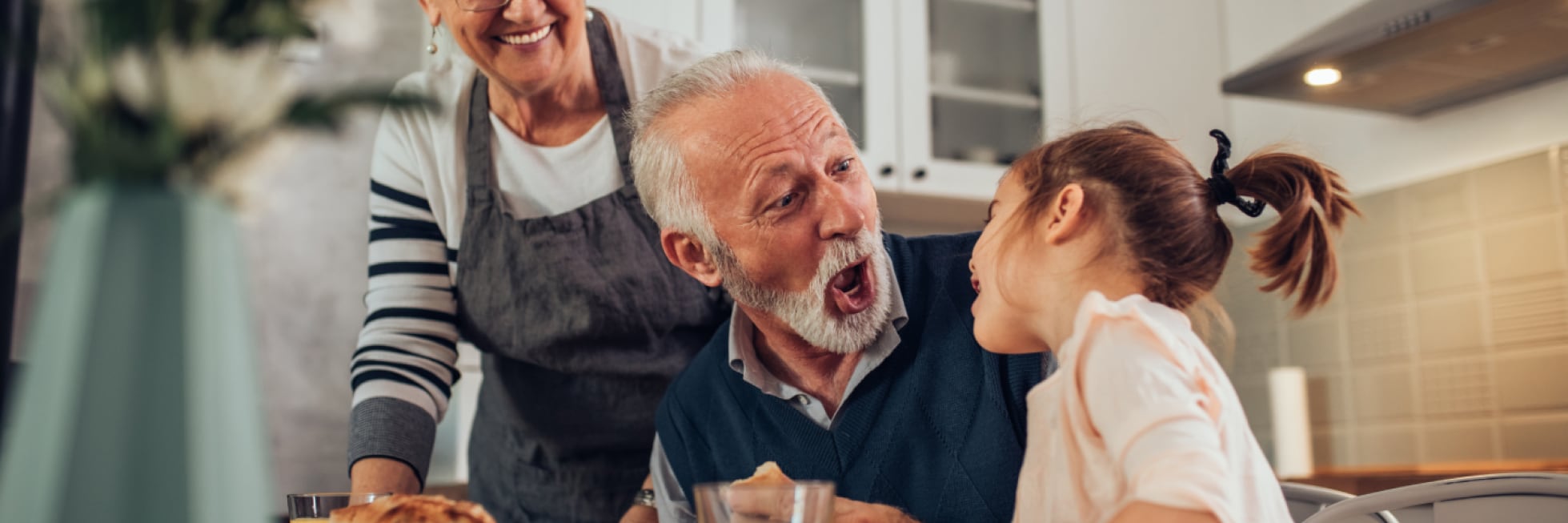 Un anciano y una niña comparten un momento divertido en una mesa de comedor, con una mujer mayor sonriendo cerca.