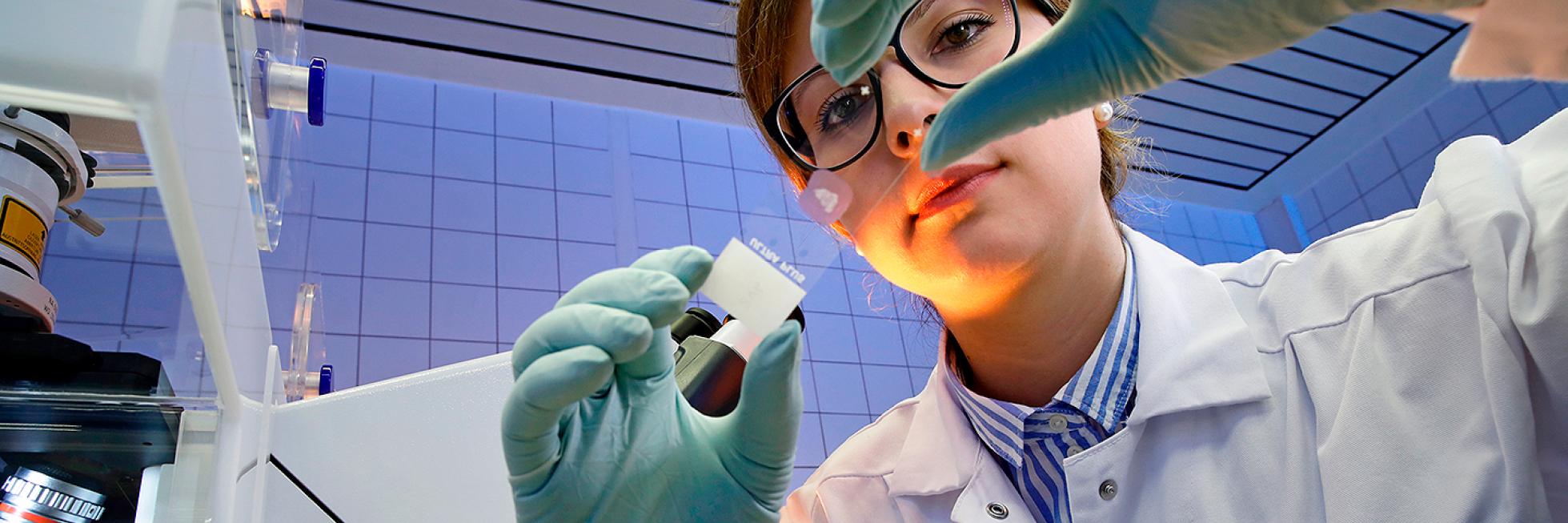 Un científico con bata y guantes examina un objeto pequeño, sosteniéndolo entre sus dedos, en un entorno de laboratorio.