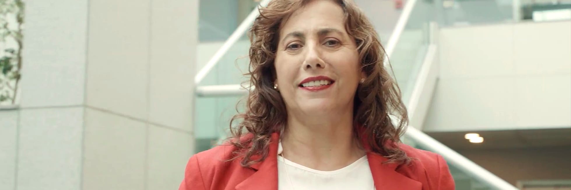 Una mujer con una chaqueta roja está de pie en el interior, sonriendo, con una escalera moderna y barandillas de cristal en el fondo.