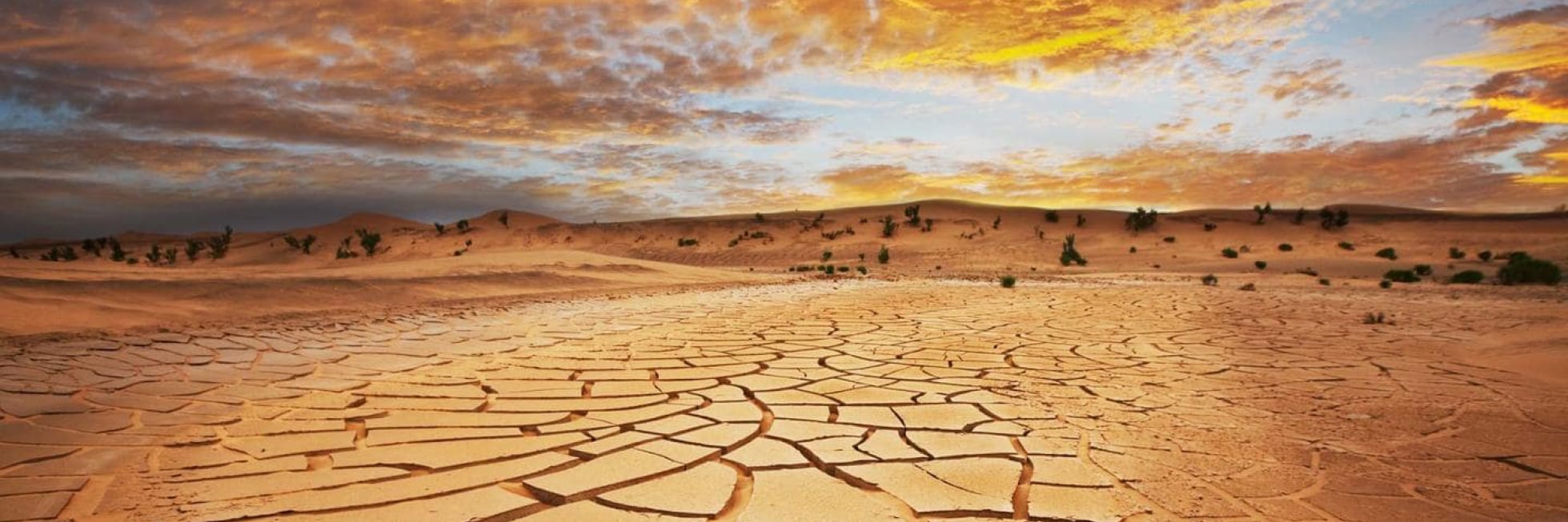 Paisaje desértico agrietado y seco bajo un espectacular cielo de atardecer con nubes dispersas y dunas distantes.