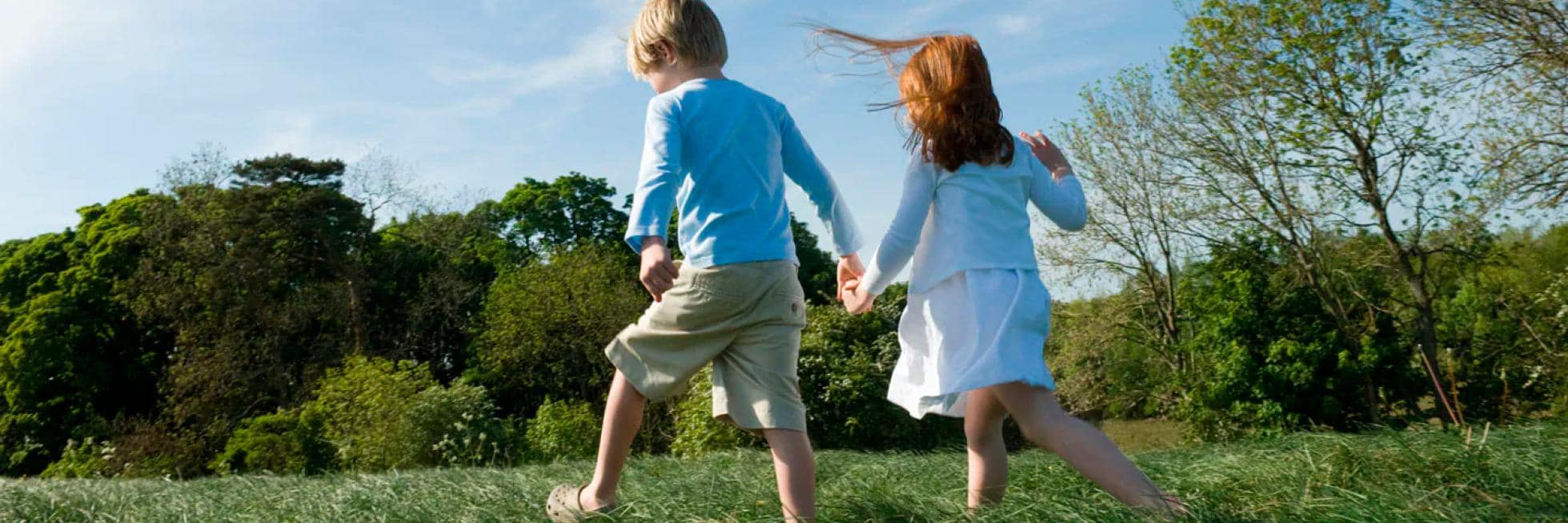 Dos niños, un niño y una niña, caminan tomados de la mano por un campo de hierba en un día soleado. Al fondo hay árboles y un cielo azul.