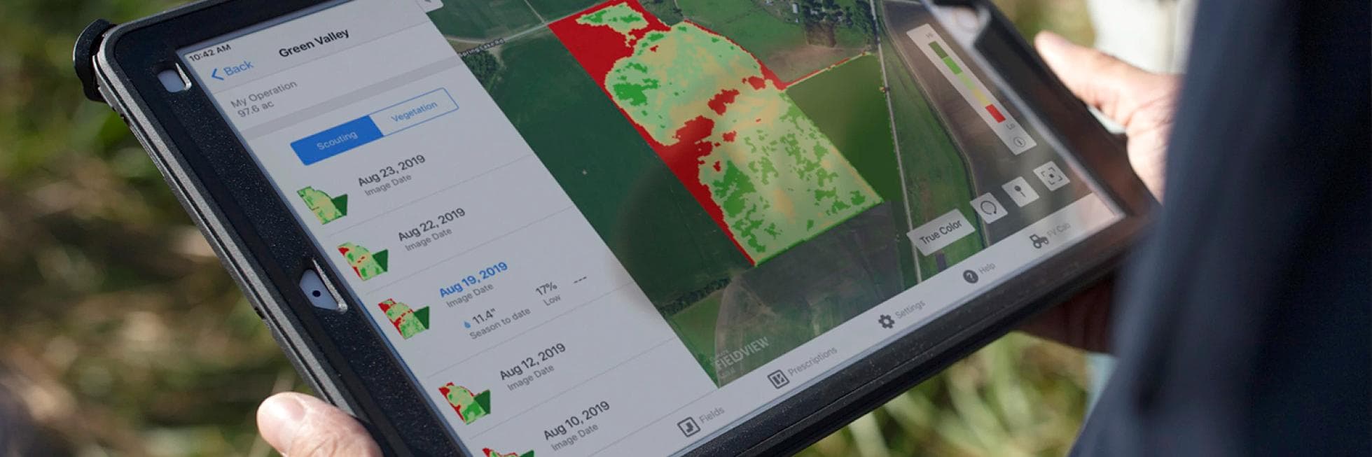 Persona que sostiene una tableta que muestra una aplicación agrícola con imágenes satelitales, mostrando áreas de campo en verde y rojo, indicando la salud del cultivo.