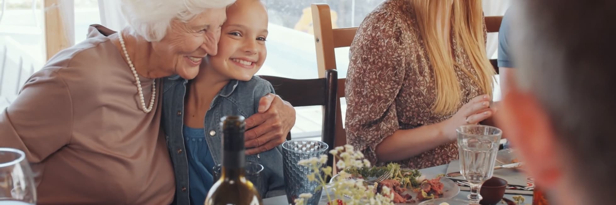 Una mujer mayor abraza a un niño sonriente en una mesa de comedor con varios platos. Otras personas también están sentadas y comiendo.