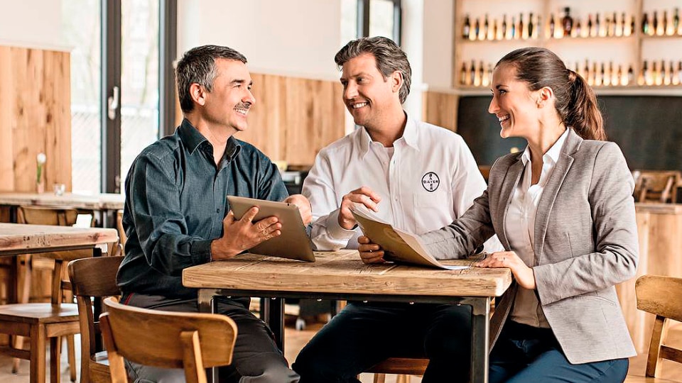 Tres personas vestidas de negocios están sentadas en una mesa en un café informal, manteniendo una conversación amistosa con tabletas.