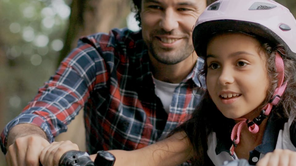 Un hombre con una camisa a cuadros ayuda a un niño que lleva un casco rosa a montar en bicicleta en un entorno forestal.