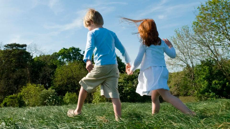 Dos niños, un niño y una niña, caminan tomados de la mano por un campo de hierba en un día soleado. Al fondo hay árboles y un cielo azul.