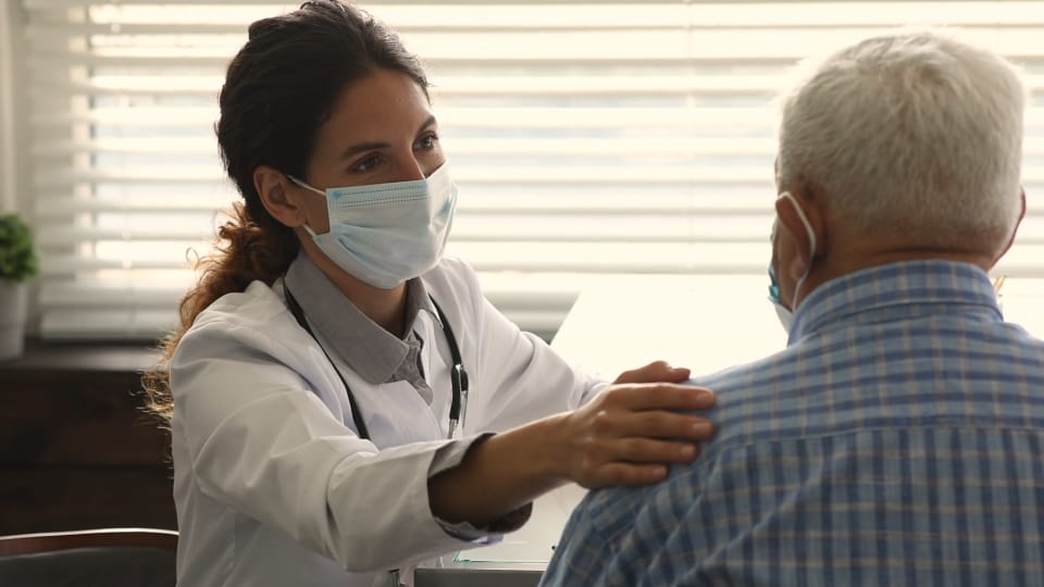 Un médico con bata blanca y mascarilla habla con un hombre mayor con mascarilla y le coloca una mano en el hombro. Detrás de ellos hay unas persianas parcialmente abiertas.