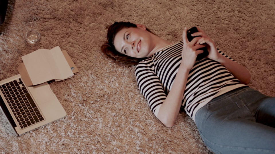 Mujer acostada sobre una alfombra, sosteniendo un teléfono, con una computadora portátil y una carpeta cerca.