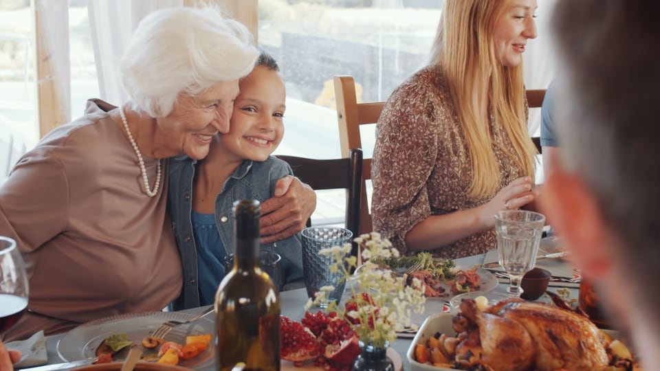 Una mujer mayor abraza a un niño sonriente en una mesa de comedor con varios platos. Otras personas también están sentadas y comiendo.