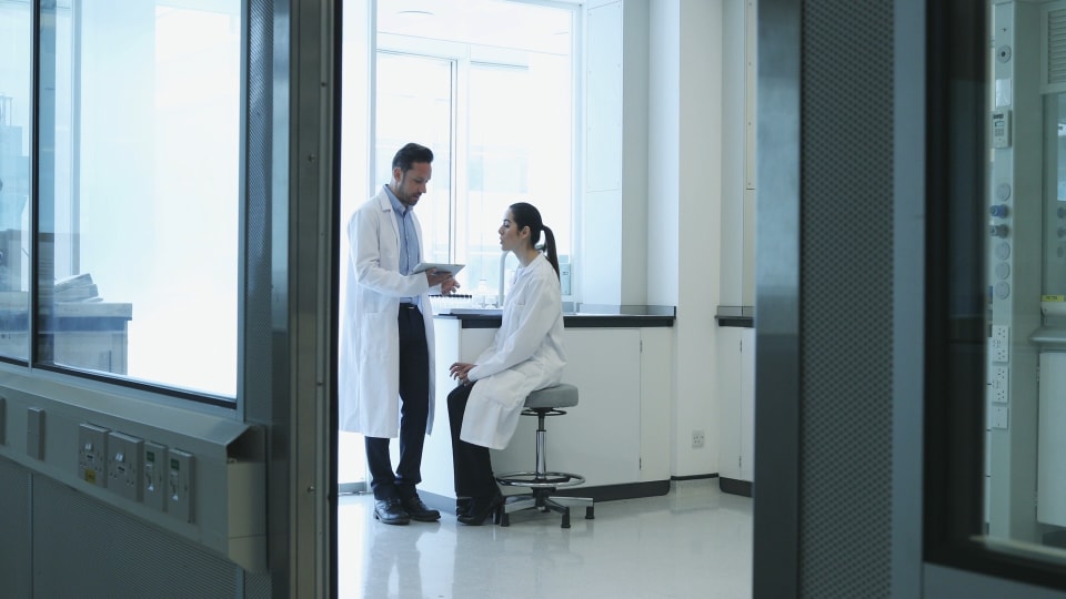 Un hombre y una mujer con bata de laboratorio conversan en una sala de laboratorio bien iluminada. La mujer está sentada en un taburete mientras el hombre está de pie sosteniendo una tableta.