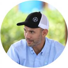 Hombre que lleva una gorra de béisbol blanca y negra y una camisa abotonada azul claro, sentado al aire libre.