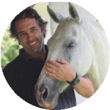 Hombre sonriendo mientras está de pie junto a un caballo blanco, apoyando suavemente su mano sobre la cara del caballo.