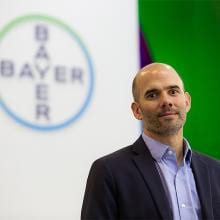 Un hombre con traje aparece delante del logotipo de Bayer sobre un fondo blanco.